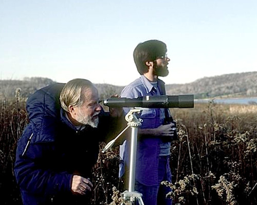 Older man looking through a telescope, while younger man looks in same direction